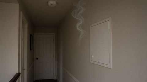 A dim hallway with a wisp of smoke near a closed wall cupboard and a smoke alarm on the ceiling